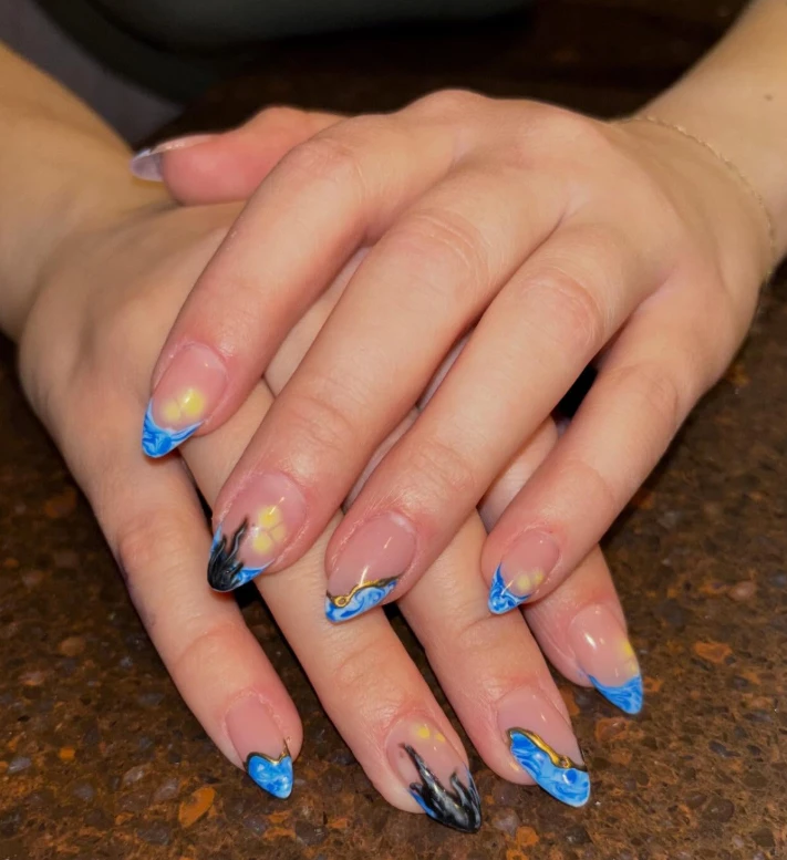 Two hands resting palm-down on a stone counter — almond-shape gel nails with blue marbled tips, citrine-yellow accents, fine gold line work, and a small hand-painted black flame on the thumb and ring fingers.